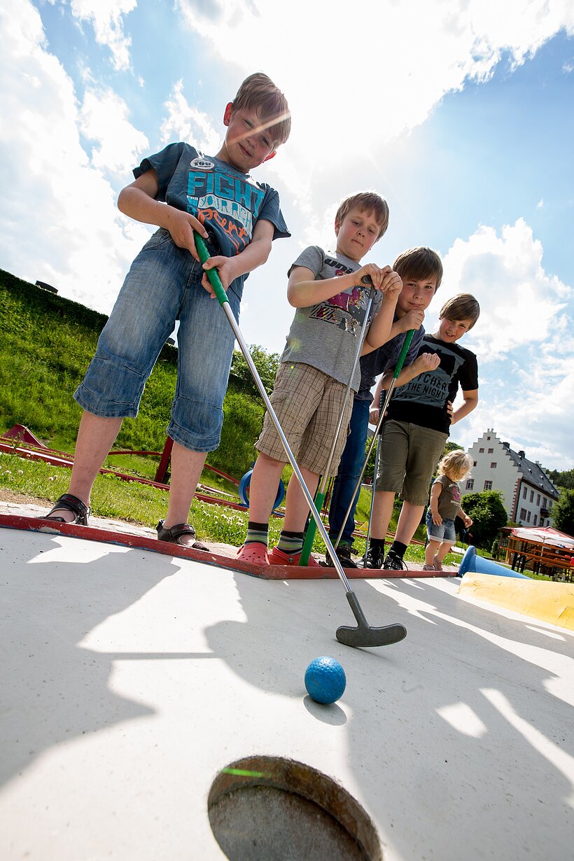 Station der Minigolfanlage mit spielenden Kindern im Vordergrund, ein Kind schiebt gerade den Ball ins Loch