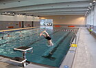 Hallenbad mit kleinem Sprungbrett am Rand und kopfüber hüpfendem Schwimmer, rechts Gehweg und Glasfensterfront