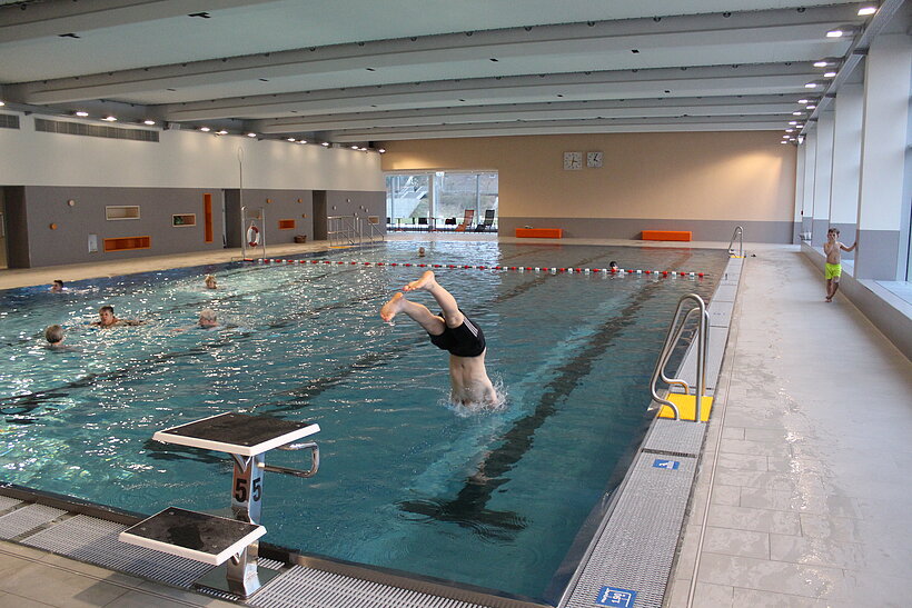 Hallenbad mit kleinem Sprungbrett am Rand und kopfüber hüpfendem Schwimmer, rechts Gehweg und Glasfensterfront