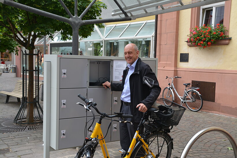 Platz mit Schließfächern, grau, Mann mit Fahrrad davor, im Hintergrund Häuserfassade mit Blumen