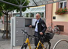 Platz mit Schließfächern, grau, Mann mit Fahrrad davor, im Hintergrund Häuserfassade mit Blumen