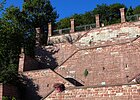 Hohe rote Sandsteinmauer, unterbrochen durch nicht sichtbare Treppe, links umringt von Bäumen und Sträuchern