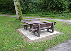 Grünfläche mit Tisch und zwei Bänken, Bäume im Hintergrund neben einem Weg