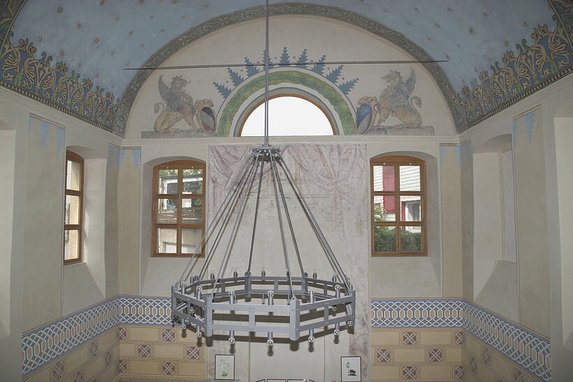 Arnstein Alte Synagoge Hellblau und olivgrüne bemalter Innenraum mit angedeuteter Kuppel, zwei Fenstern und einem Halbrundfenster darüber, in der Mitte hängt ein großer Leuchter