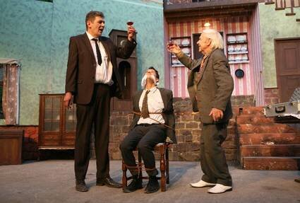 Szene eines Theaterstücks, ein Mann sitzt auf einem Stuhl zwischen zwei Männern, die sich zuprosten