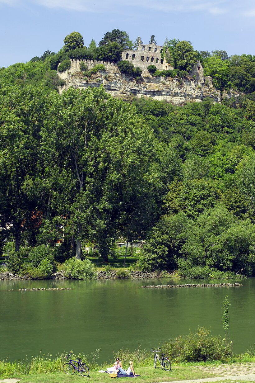 Fluss vorne, dahinter Anhöhe mit viel Grün und Burgruine oben