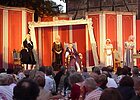 Freiluftbühne: rotgoldene Kulisse mit barockgewandeten Schauspielern, Zuschauer davor