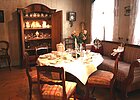 Raum mit zwei Fenstern mit hellen Vorhängen rechts, an der Wand im Hintergrund ein geöffneter Holzschrank mit Regalen darin, dort stehen Ausstellungsstücke, in der Mitte des Raumes ein runder Tisch mit antiken Stühlen
