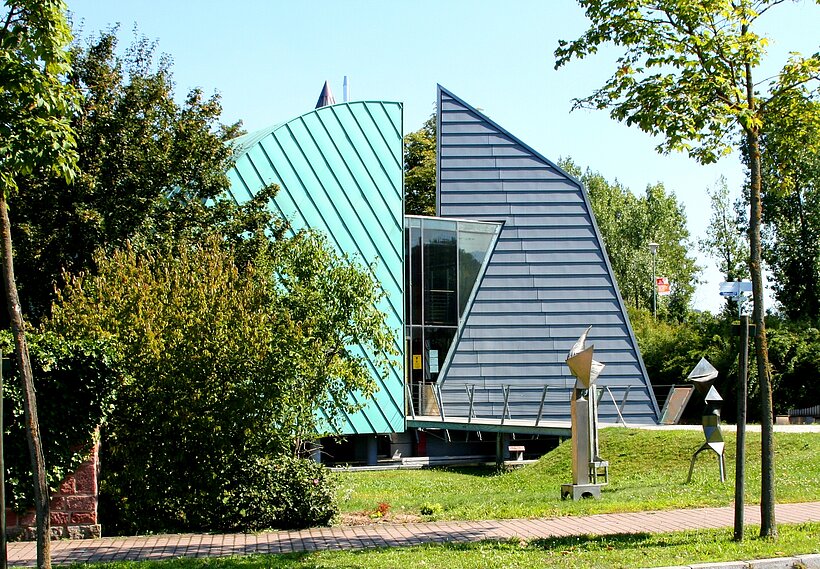 Klempnermuseum Karlstadt Ungewöhnliches Haus im Freien, bestehend aus zwei hohen geriffelten ungleichen Metallwänden in hell und dunkelblau. Es steht inmitten von Grün und Bäumen