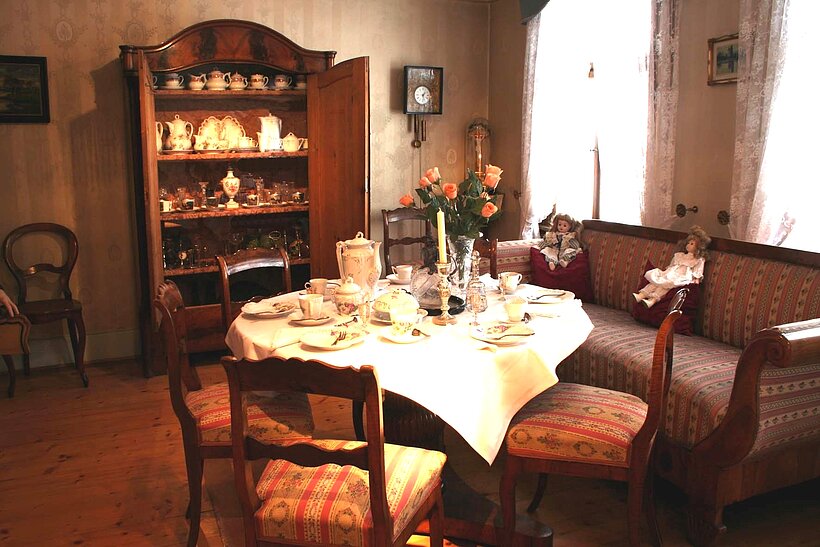 Heimatmuseum Karlstadt Raum mit zwei Fenstern mit hellen Vorhängen rechts, an der Wand im Hintergrund ein geöffneter Holzschrank mit Regalen darin, dort stehen Ausstellungsstücke, in der Mitte des Raumes ein runder Tisch mit antiken Stühlen