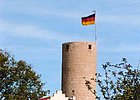 Weiße Giebelfassade mit kleinen Zinnen, Steinturm mit Fahne daneben, rechts und links zwei Bäume